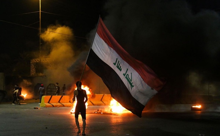 متظاهر في مدينة كربلاء يحمل العلم العراقي أمام النيران المشتعلة عند بوابة القنصلية الإيرانية في المدينة
